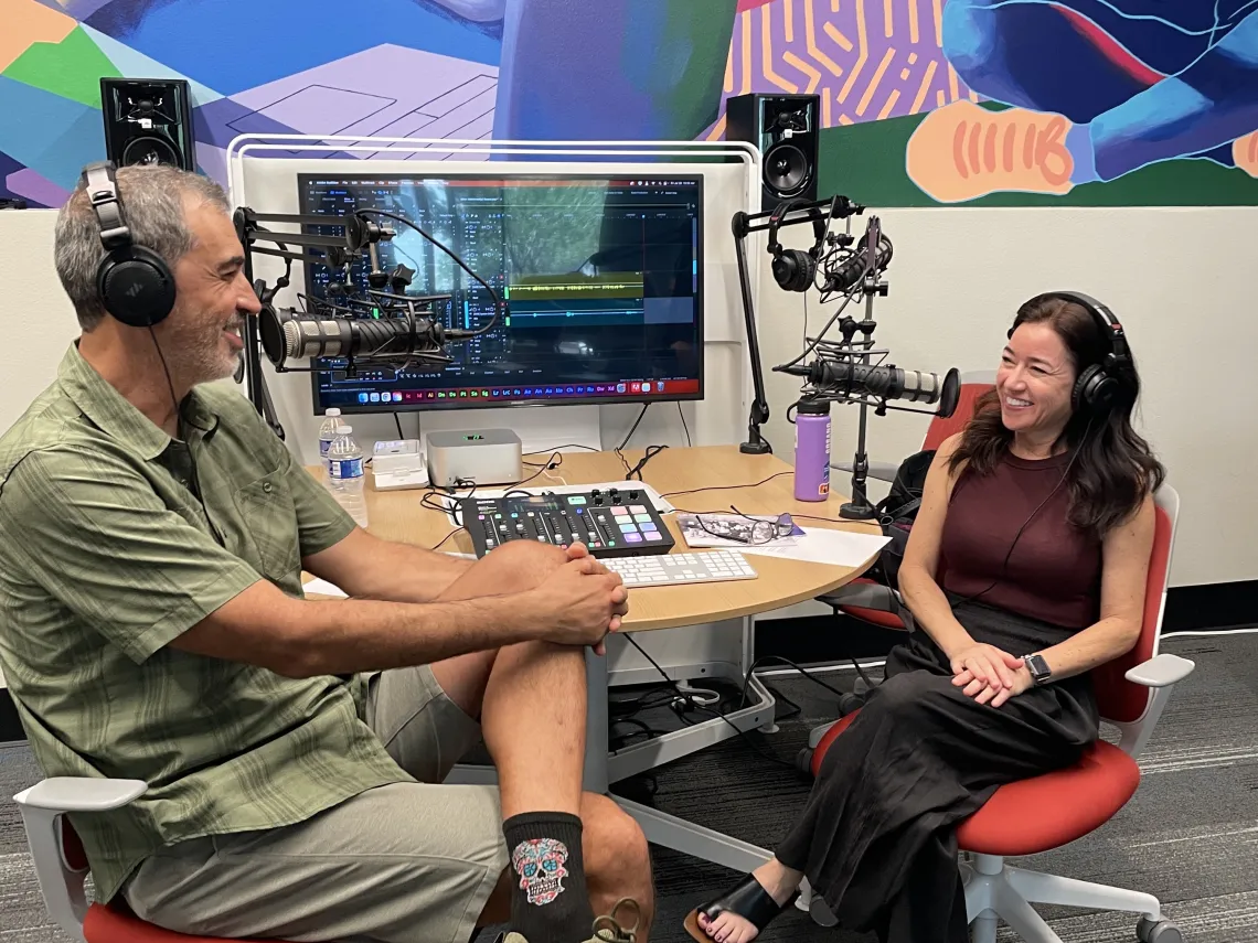 Two people sitting in front of tv screen wearing headphones speaking into microphones