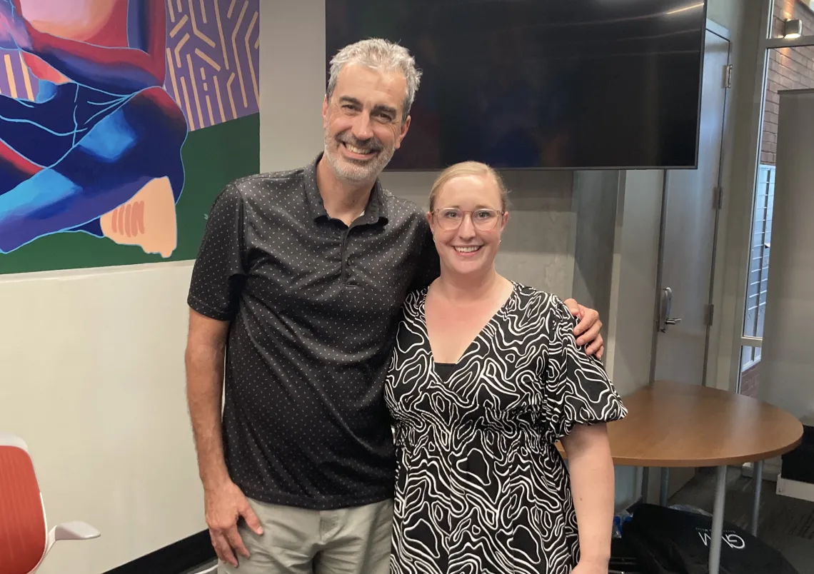 John Pollard and Jacqueline Melvold standing in podcast studio smiling for camera