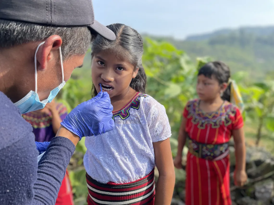 A young girl in Guatemala looks at the camera while getting her teeth checked by a healthcare worker