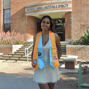 graduate standing in front of brick building wearing yellow and white stole