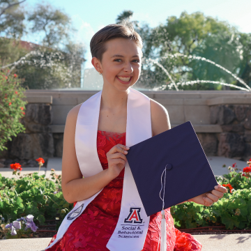 graduate wearing red dress and white stole sitting in front of fountain holding blue cap 